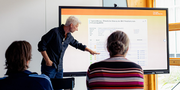 Ein Mann mit weißen Haaren steht vor einem Smartboard und erklärt einer Gruppe von Teilnehmenden das Planspiel