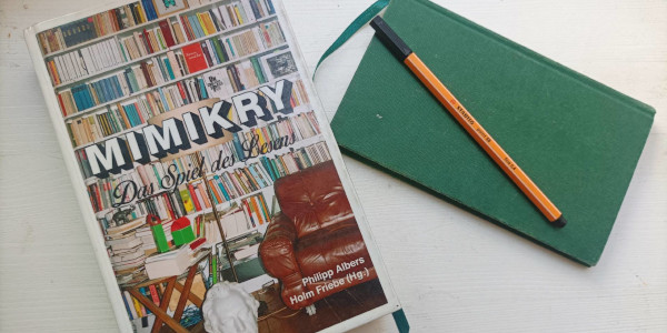 Bild von einem Buch auf dem ein großes Bücherregal zu sehen ist und daneben ein Notizbuch mit Stift