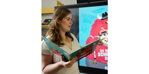 Eine Frau mit braunen Haaren liest aus einem Bilderbuch vor und zeigt auf einem Bildschirm ein Bilderbuchkino
