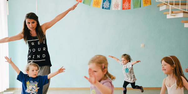 Eine Frau und vier Kinder tanzen mit ausgestreckten Armen wie ein Flugzeug. Im Hintergrund ist eine Girlande zu sehen.