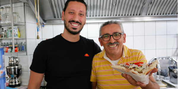 ein älterer Mann streckt einen Döner Richtung Kamera und lächelt, neben ihm steht ein jüngerer Mann und lächelt 