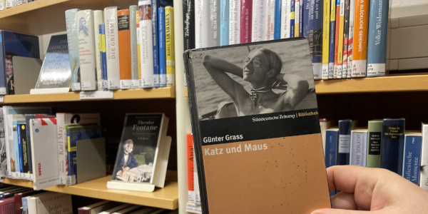 Eine Person hält eine Ausgabe von Günter Grass' „Katz und Maus“ vor einem Bücherregal in der Bibliothek
