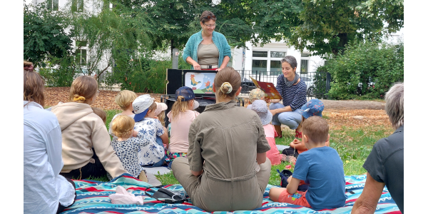 Zwei Bibliotheksmitarbeiterinnen führen ein Kamishibai-Theater auf einer Wiese auf, im Publikum sind viele Kinder.