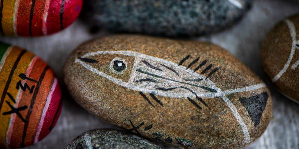 Foto eines angemalten Steines mit fisch drauf. Darum liegen angedeutet weitere bunt bemalte Steine..